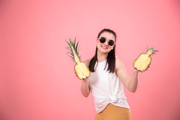 Stylish young girl in sunglasses smiles and holds fruits on a pink background. Summer vacation concept.