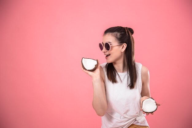 Stylish young girl in sunglasses smiles and holds fruits on a pink background. Summer vacation concept.