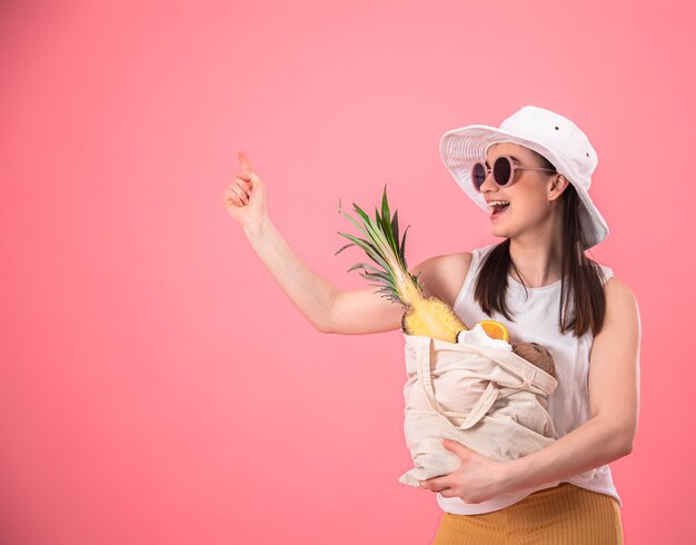 Стильная молодая девушка в белой шляпе и солнечных очках улыбается и держит эко-сумку с экзотическими фруктами