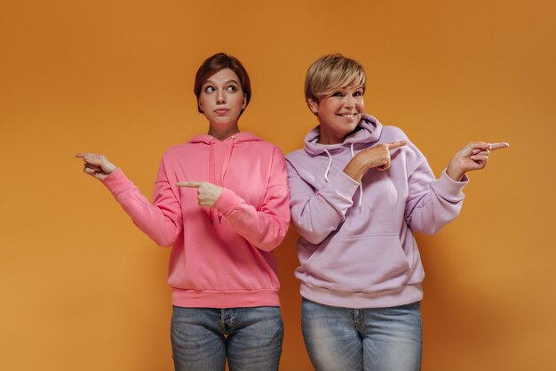 Stylish young girl with brunette hair in pink sweater pointing fingers to side together with old lady with modern outfit on orange backdrop. 