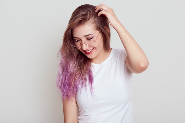 Stylish young girl with lilac hair wearing white t shirt and glasses with positive expression, looking down, keeping hand on her head, dreams, posing isolated over gray wall.