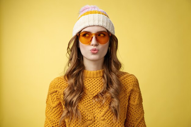 スタイリッシュな若い魅力思慮深い女の子は、休暇のスキーリゾート、黄色の背景を楽しんでいるサングラス帽子暖かいセーターを着て左に目をそらす製品を選ぶことを考えて愚かな唇を折りたたむ乾燥を作ります