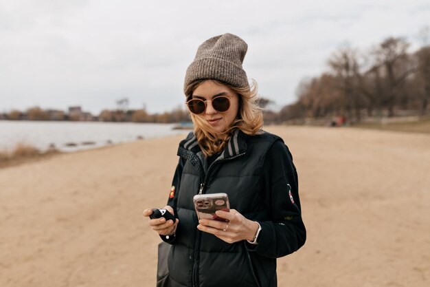 선글라스와 모자를 쓴 세련된 젊은 여성이 화창한 봄날 해변에서 스마트폰을 스크롤하고 있습니다
