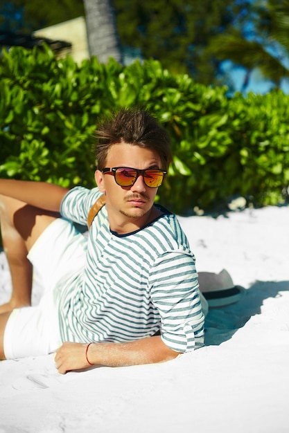 Stylish young male model man lying on beach sand wearing hipster summer hat enjoying summer travel holiday near ocean in sunglasses