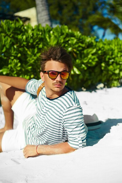 Stylish young male model man lying on beach sand  wearing hipster summer hat  enjoying summer travel holiday near ocean in sunglasses