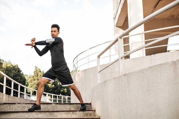 검은 색 스포츠 반바지와 긴팔 티셔츠에 세련된 젊은 남자가 뻗어 계단에서 야외에서 운동합니다.