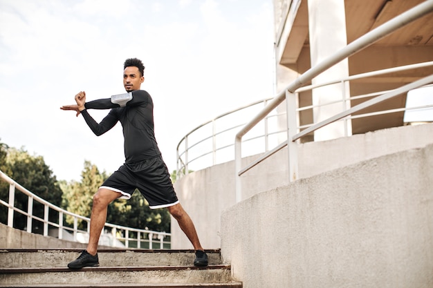 Stylish young man in black sport shorts and long-sleeved t-shirt stretches and works out outdoors on stairs