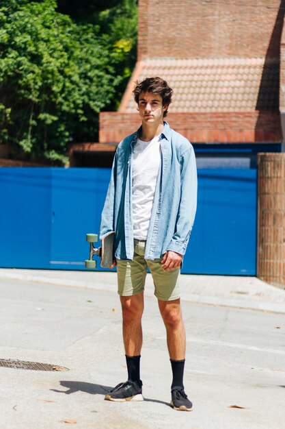 Stylish young man holding skateboard standing outside