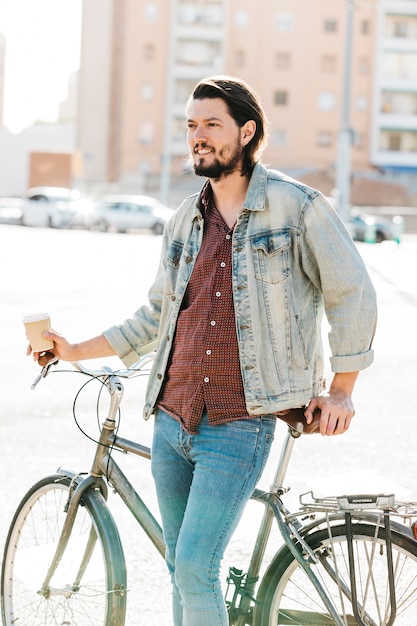 離れている紙のコーヒーカップを保持している自転車にもたれてスタイリッシュな若い男