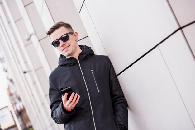 Стильный молодой человек, опираясь на стену, держа в руке мобильный телефон