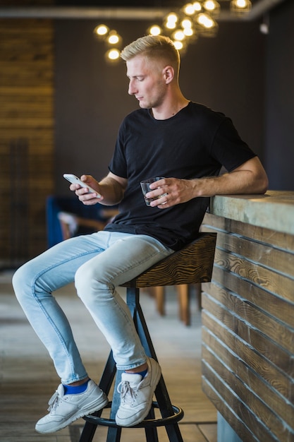 Stylish young man sitting on bar stool holding drink using smartphone