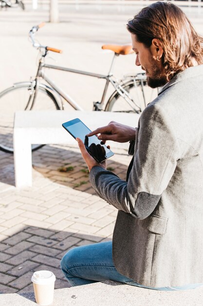 携帯電話を使用してテイクアウトのコーヒーカップとベンチに座っているスタイリッシュな若い男