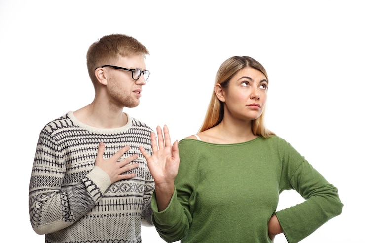Free Photo stylish young man in spectacles holding hand on his chest, asking his angry girlfriend for forgiveness while woman not looking at him, making stop gesture as if saying: i don't need your excuse