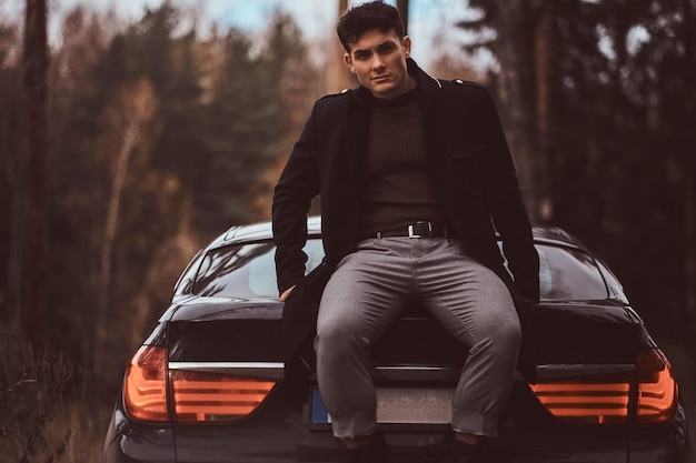 Free photo a stylish young man wearing a black coat sitting on the trunk of a car in the autumn forest in the evening.