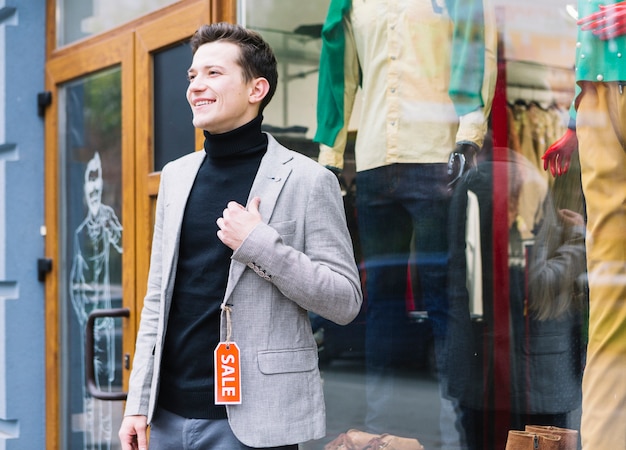 Stylish young man wearing jacket with sale tag standing outside the shop