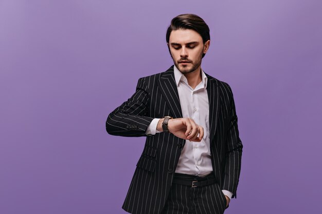 ブルネットの髪、白いシャツ、紫色の壁に隔離された彼の手で時計を見ている古典的な縞模様のスーツを持つスタイリッシュな若い男