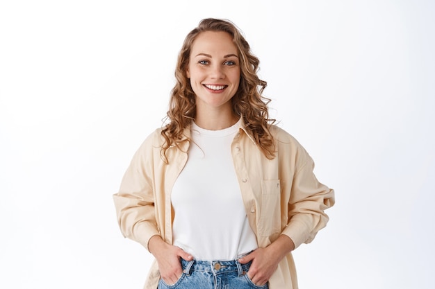 Бесплатное фото Стильная молодая современная женщина со светлой прической, держится за руки в карманах джинсов и беззаботно улыбается, выглядит уверенно и решительно, белая стена