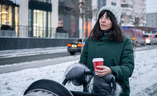 Stylish young mom with a cup of coffee on a walk with a baby carriage in winter