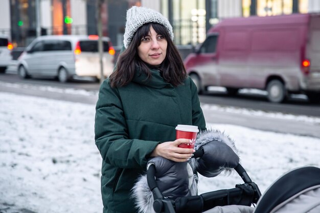 겨울에 유모차를 타고 산책하며 커피 한 잔을 들고 있는 세련된 젊은 엄마