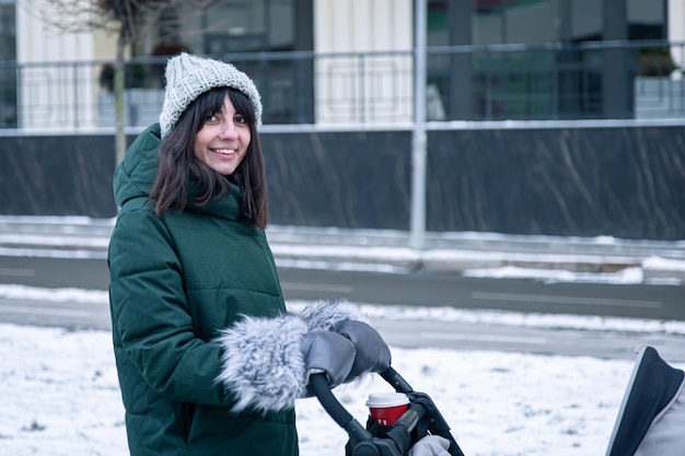 Free photo stylish young mom with a cup of coffee on a walk with a baby carriage in winter