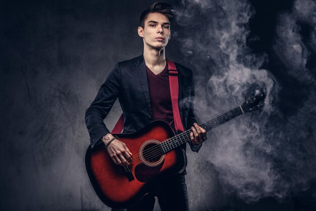 Stylish young musician with stylish hair in elegant clothes with a guitar in his hands playing and posing on a dark background.