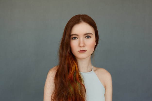 stylish young redhead Caucasian female model with multicolored strands of hair posing at blank wall, looking with calm expression on her face. Style and fashion concept