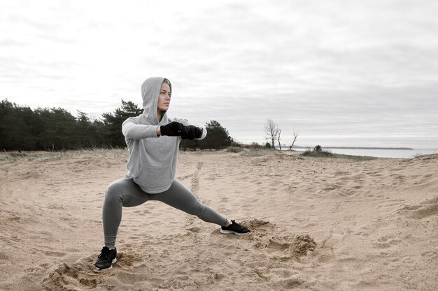 Stylish young sportswoman wearing gloves, hoodie and sneakers doing standing yoga pose for strong legs, preparing muscles for cardio workout. Confident athletic female in hood exercising on beach