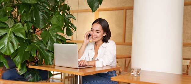 Free Photo stylish young woman answer phone call sitting in cafe with laptop working and taling on smartphone