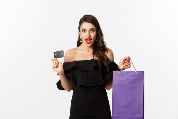 Stylish young woman in black dress going shopping, holding bag and credit card, smiling pleased, standing over white background.