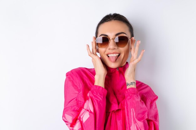 Стильная молодая женщина в ярко-розовой спортивной куртке, модных солнцезащитных очках и беспроводных наушниках в ушах на белом фоне показывает язык