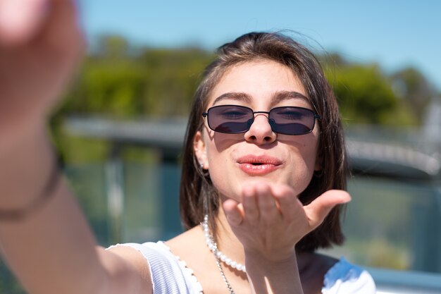 Стильная молодая женщина в повседневном белом кроп-топе и солнечных очках делает селфи на мобильном телефоне, веселые, возбужденные, позитивные флюиды
