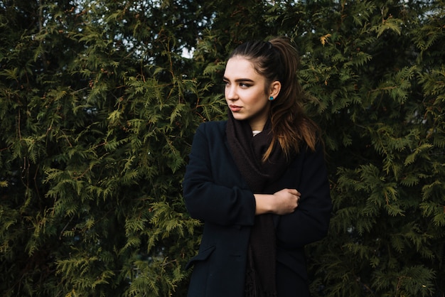 Stylish young woman in coat with scarf near coniferous plants