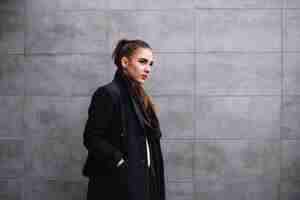 Free photo stylish young woman in coat with scarf near grey wall