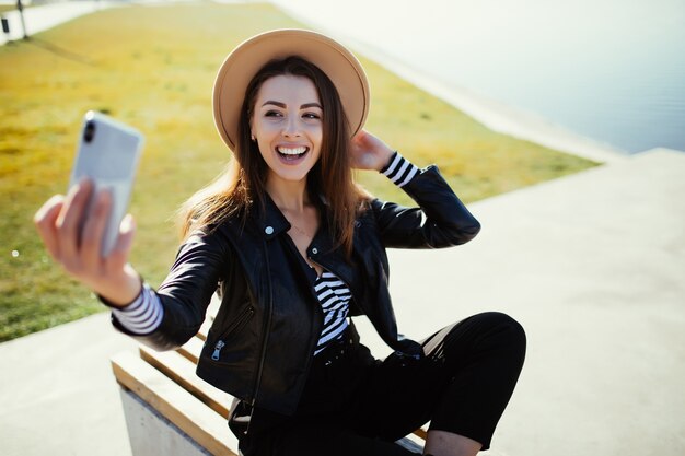 Stylish young woman girl make selfie in the park near the city lake in cold sunny summer day dressed up in black clothes