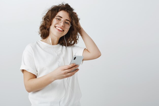 Stylish young woman in glasses making playlist in phone, listening music via earphones