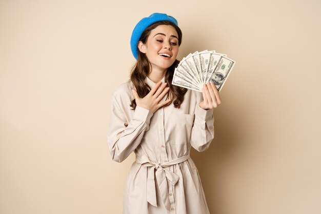 돈 현금 달러를 들고 웃는 세련된 젊은 여성이 쇼핑 스탠드에 만족스럽게 포즈를 취하고 있습니다.