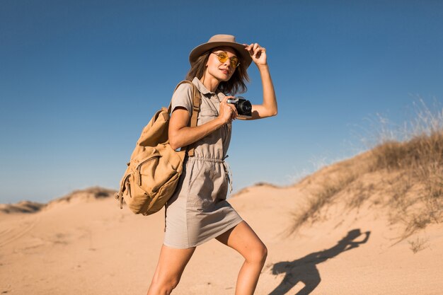 Стильная молодая женщина в платье цвета хаки гуляет по песку пустыни, путешествует по Африке на сафари, в шляпе и рюкзаке, фотографирует на старинную камеру