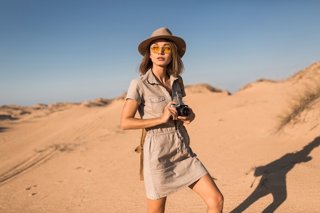砂漠を歩いて、サファリでアフリカを旅行し、帽子とバックパックを身に着けて、ビンテージカメラで写真を撮るカーキ色のドレスを着たスタイリッシュな若い女性