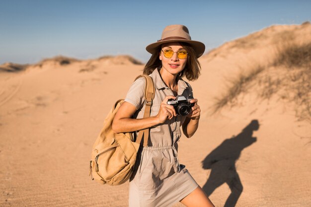 Stylish young woman in khaki dress walking in desert, traveling in Africa on safari, wearing hat and backpack, taking photo on vintage camera