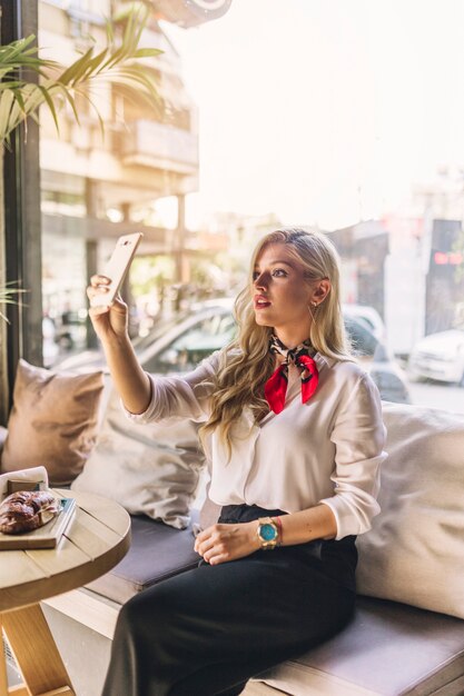 Стильная молодая женщина сидит в кафе, принимая селфи со смартфона