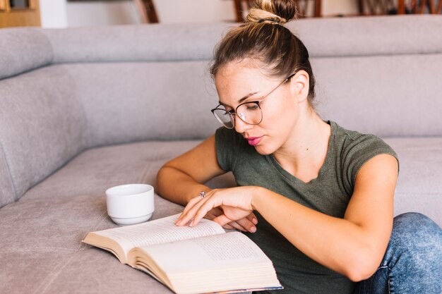 Stylish young woman sitting near the sofa reading book