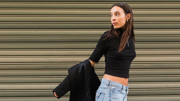 Stylish young woman standing in front of iron shutter looking over shoulder removing the jacket