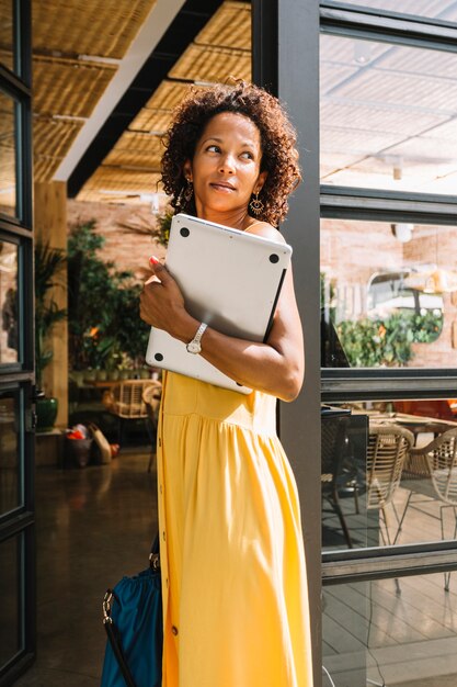 Stylish young woman standing near the restaurant entrance holding laptop