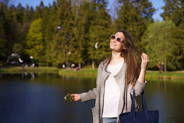 バッグとサングラスを着たスタイリッシュな若い女性が彼女の手に花を晴れた日に湖を背景に歩く