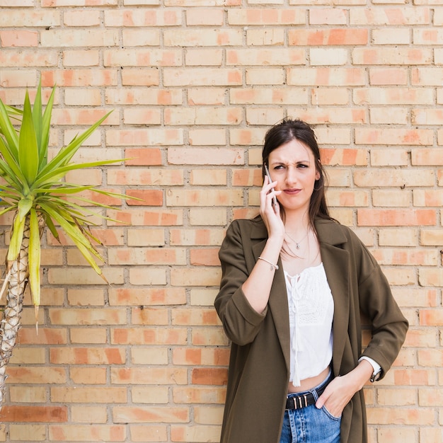 Free photo stylish young woman talking on mobile phone standing against brick wall