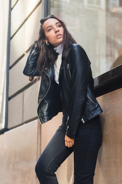 Stylish young woman wearing black jacket leaning on wall