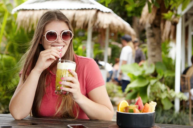 スタイリッシュな若い女性がバーのカウンターに座って丸い色合いを着てストローでフルーツシェイクを飲みながら、リラックスして暑いエキゾチックな国での休暇中に晴れた日を楽しんでいます。