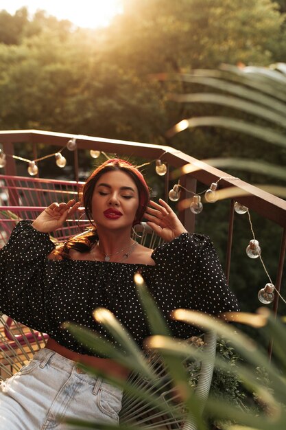 Stylish young woman with bandana and earrings in black polka dot blouse and jeans sitting and posing with closed eyes on terrace