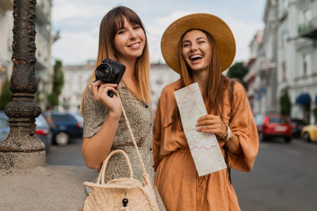 春の流行のドレスやアクセサリーを身に着けて一緒に旅行するスタイリッシュな若い女性は、地図を保持しているカメラで写真を撮ることを楽しんでいます