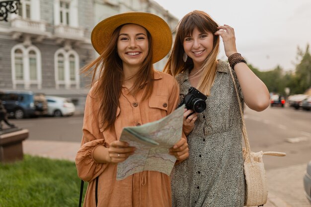 Stylish young women travelling together in Europe dressed in spring trendy dresses and accessories holding map